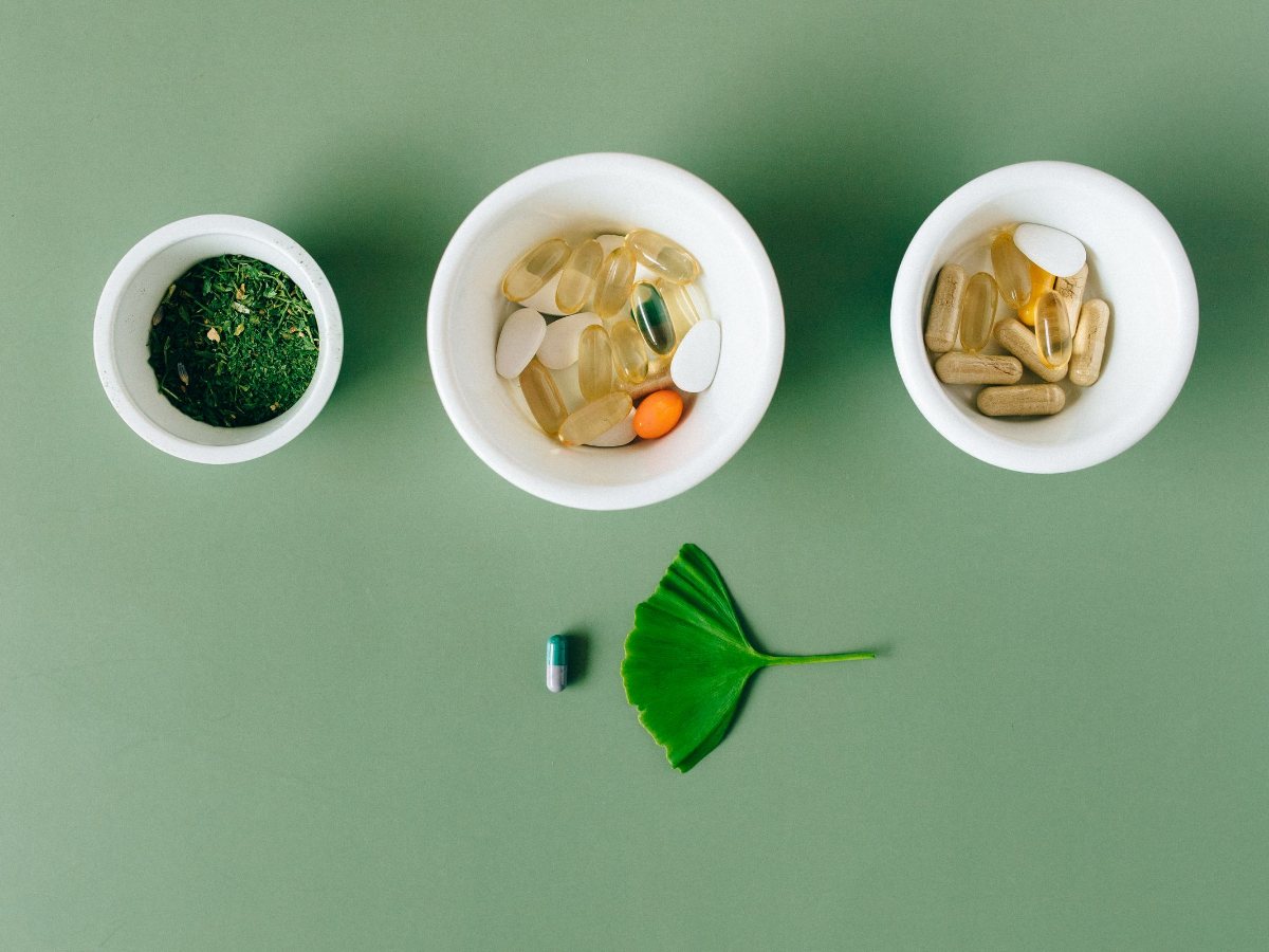 Capsules, tablets and dried herbs in white bowls on a sage-green surface